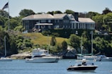 A large, stately mansion with classical architecture sits atop a grassy hill overlooking a body of water. The mansion is constructed with stone and has a number of tall, white columns at one end. Lush green trees and vegetation surround the property. Several boats, including yachts and smaller motorboats, are anchored in the water near the shoreline. An American flag is visible on a flagpole near the mansion.