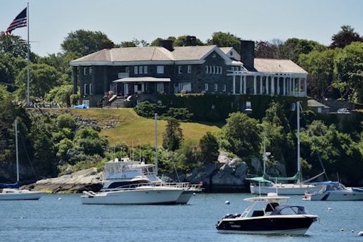 A large, stately mansion with classical architecture sits atop a grassy hill overlooking a body of water. The mansion is constructed with stone and has a number of tall, white columns at one end. Lush green trees and vegetation surround the property. Several boats, including yachts and smaller motorboats, are anchored in the water near the shoreline. An American flag is visible on a flagpole near the mansion.