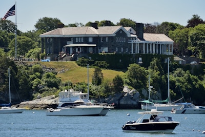 A large, stately mansion with classical architecture sits atop a grassy hill overlooking a body of water. The mansion is constructed with stone and has a number of tall, white columns at one end. Lush green trees and vegetation surround the property. Several boats, including yachts and smaller motorboats, are anchored in the water near the shoreline. An American flag is visible on a flagpole near the mansion.