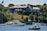 A large, stately mansion with classical architecture sits atop a grassy hill overlooking a body of water. The mansion is constructed with stone and has a number of tall, white columns at one end. Lush green trees and vegetation surround the property. Several boats, including yachts and smaller motorboats, are anchored in the water near the shoreline. An American flag is visible on a flagpole near the mansion.
