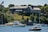 A large, stately mansion with classical architecture sits atop a grassy hill overlooking a body of water. The mansion is constructed with stone and has a number of tall, white columns at one end. Lush green trees and vegetation surround the property. Several boats, including yachts and smaller motorboats, are anchored in the water near the shoreline. An American flag is visible on a flagpole near the mansion.