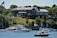 A large, stately mansion with classical architecture sits atop a grassy hill overlooking a body of water. The mansion is constructed with stone and has a number of tall, white columns at one end. Lush green trees and vegetation surround the property. Several boats, including yachts and smaller motorboats, are anchored in the water near the shoreline. An American flag is visible on a flagpole near the mansion.