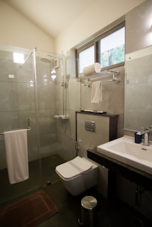 A modern bathroom with a glass shower enclosure featuring metal fixtures and a mounted showerhead. There is a wall-mounted toilet underneath a towel rack holding folded towels. A sink with a rectangular basin is positioned next to the toilet, and a small trash bin is on the floor beside it. The bathroom has tiled walls and a dark floor mat, and a window provides natural light.