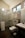 A modern bathroom with a glass shower enclosure featuring metal fixtures and a mounted showerhead. There is a wall-mounted toilet underneath a towel rack holding folded towels. A sink with a rectangular basin is positioned next to the toilet, and a small trash bin is on the floor beside it. The bathroom has tiled walls and a dark floor mat, and a window provides natural light.