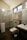 A modern bathroom with a glass shower enclosure featuring metal fixtures and a mounted showerhead. There is a wall-mounted toilet underneath a towel rack holding folded towels. A sink with a rectangular basin is positioned next to the toilet, and a small trash bin is on the floor beside it. The bathroom has tiled walls and a dark floor mat, and a window provides natural light.