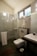 A modern bathroom with a glass shower enclosure featuring metal fixtures and a mounted showerhead. There is a wall-mounted toilet underneath a towel rack holding folded towels. A sink with a rectangular basin is positioned next to the toilet, and a small trash bin is on the floor beside it. The bathroom has tiled walls and a dark floor mat, and a window provides natural light.
