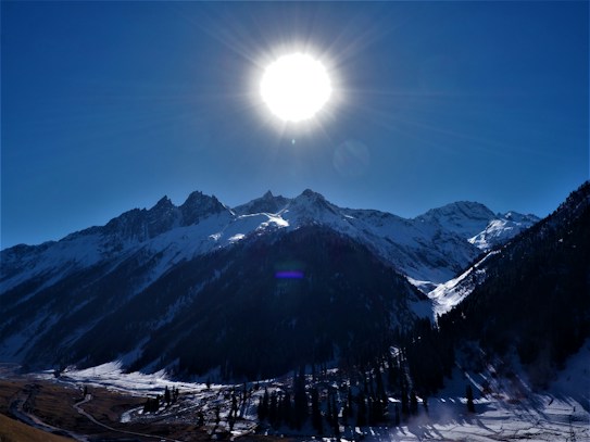 The image captures a stunning view of snow-capped mountains under a bright sun, creating a dramatic sky with lens flare effects. Pine trees dot the landscape at the base of the mountains, while a winding stream carves through the snow-covered valley beneath.