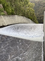 A powerful flow of water is cascading down a smooth, wide concrete structure, likely a dam spillway. Surrounding the spillway are lush green trees and dense forested hills.