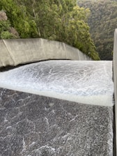 A powerful flow of water is cascading down a smooth, wide concrete structure, likely a dam spillway. Surrounding the spillway are lush green trees and dense forested hills.