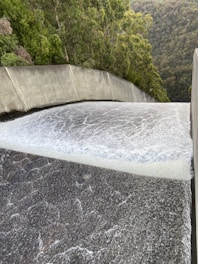 A powerful flow of water is cascading down a smooth, wide concrete structure, likely a dam spillway. Surrounding the spillway are lush green trees and dense forested hills.