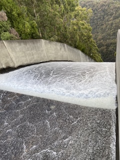 A powerful flow of water is cascading down a smooth, wide concrete structure, likely a dam spillway. Surrounding the spillway are lush green trees and dense forested hills.