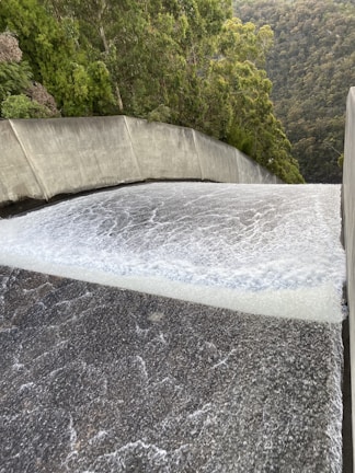 A powerful flow of water is cascading down a smooth, wide concrete structure, likely a dam spillway. Surrounding the spillway are lush green trees and dense forested hills.