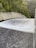A powerful flow of water is cascading down a smooth, wide concrete structure, likely a dam spillway. Surrounding the spillway are lush green trees and dense forested hills.