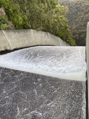 A powerful flow of water is cascading down a smooth, wide concrete structure, likely a dam spillway. Surrounding the spillway are lush green trees and dense forested hills.