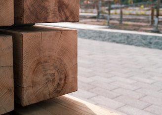 Several neatly stacked wooden beams with visible growth rings are seen in the foreground, showcasing their natural texture and grain. The background is an outdoor space with a paved area and some blurred elements, likely trees or fencing.
