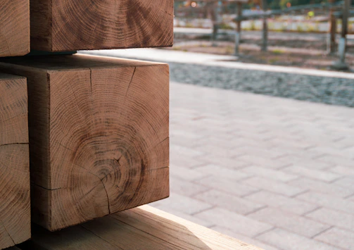 Several neatly stacked wooden beams with visible growth rings are seen in the foreground, showcasing their natural texture and grain. The background is an outdoor space with a paved area and some blurred elements, likely trees or fencing.