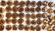 A neatly arranged grid of chocolate chip cookies with varying toppings, including chocolate drizzle, small chocolate balls, and white marshmallows. The cookies have a mix of brown and chocolatey hues, creating a visually tempting display.