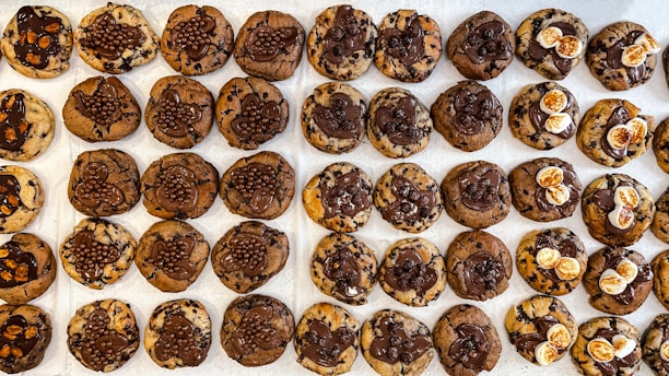 A neatly arranged grid of chocolate chip cookies with varying toppings, including chocolate drizzle, small chocolate balls, and white marshmallows. The cookies have a mix of brown and chocolatey hues, creating a visually tempting display.