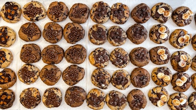 A neatly arranged grid of chocolate chip cookies with varying toppings, including chocolate drizzle, small chocolate balls, and white marshmallows. The cookies have a mix of brown and chocolatey hues, creating a visually tempting display.