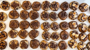 A neatly arranged grid of chocolate chip cookies with varying toppings, including chocolate drizzle, small chocolate balls, and white marshmallows. The cookies have a mix of brown and chocolatey hues, creating a visually tempting display.