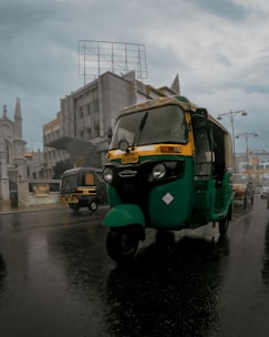 A green auto rickshaw drives down a wet city street under overcast skies. Buildings with modern and traditional architecture flank the road while other vehicles and pedestrians are visible in the background.