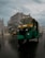 A green auto rickshaw drives down a wet city street under overcast skies. Buildings with modern and traditional architecture flank the road while other vehicles and pedestrians are visible in the background.