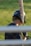 A person wearing a baseball helmet and batting gloves is holding a baseball bat over their shoulder. The bat has the brand name 'Marucci' visible on it. The background is a grassy area, and the image is partially obstructed by horizontal bars in the foreground.