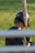 A person wearing a baseball helmet and batting gloves is holding a baseball bat over their shoulder. The bat has the brand name 'Marucci' visible on it. The background is a grassy area, and the image is partially obstructed by horizontal bars in the foreground.