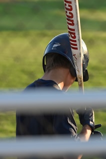 A person wearing a baseball helmet and batting gloves is holding a baseball bat over their shoulder. The bat has the brand name 'Marucci' visible on it. The background is a grassy area, and the image is partially obstructed by horizontal bars in the foreground.