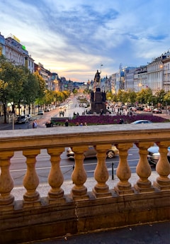 A bustling city street lined with historic buildings is visible from behind a stone balustrade. In the center, a large equestrian statue stands surrounded by flowers. The busy street is filled with people and cars, with the sky painted in shades of blue and orange as the sun sets.
