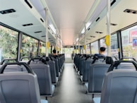The interior of a city bus with rows of empty seats, except for two passengers seated towards the back. Large windows on each side provide a view of greenery and city buildings outside. Overhead handrails and straps are available for standing passengers. The bus is clean and well-lit, with a modern design.