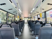 The interior of a city bus with rows of empty seats, except for two passengers seated towards the back. Large windows on each side provide a view of greenery and city buildings outside. Overhead handrails and straps are available for standing passengers. The bus is clean and well-lit, with a modern design.