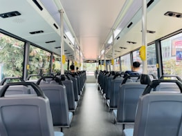 The interior of a city bus with rows of empty seats, except for two passengers seated towards the back. Large windows on each side provide a view of greenery and city buildings outside. Overhead handrails and straps are available for standing passengers. The bus is clean and well-lit, with a modern design.