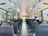 The interior of a city bus with rows of empty seats, except for two passengers seated towards the back. Large windows on each side provide a view of greenery and city buildings outside. Overhead handrails and straps are available for standing passengers. The bus is clean and well-lit, with a modern design.