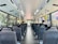 The interior of a city bus with rows of empty seats, except for two passengers seated towards the back. Large windows on each side provide a view of greenery and city buildings outside. Overhead handrails and straps are available for standing passengers. The bus is clean and well-lit, with a modern design.