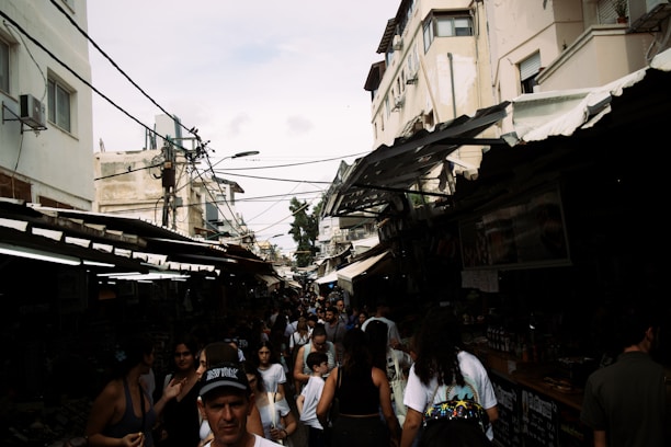 A bustling outdoor market with many people walking between tightly packed stalls. The market is situated in a narrow alleyway between buildings with weathered exteriors. Overhead wires crisscross above the scene, while a mix of sunlight and shadows creates a dynamic and slightly chaotic atmosphere.