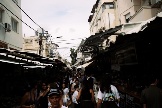 A bustling outdoor market with many people walking between tightly packed stalls. The market is situated in a narrow alleyway between buildings with weathered exteriors. Overhead wires crisscross above the scene, while a mix of sunlight and shadows creates a dynamic and slightly chaotic atmosphere.