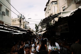 A bustling outdoor market with many people walking between tightly packed stalls. The market is situated in a narrow alleyway between buildings with weathered exteriors. Overhead wires crisscross above the scene, while a mix of sunlight and shadows creates a dynamic and slightly chaotic atmosphere.