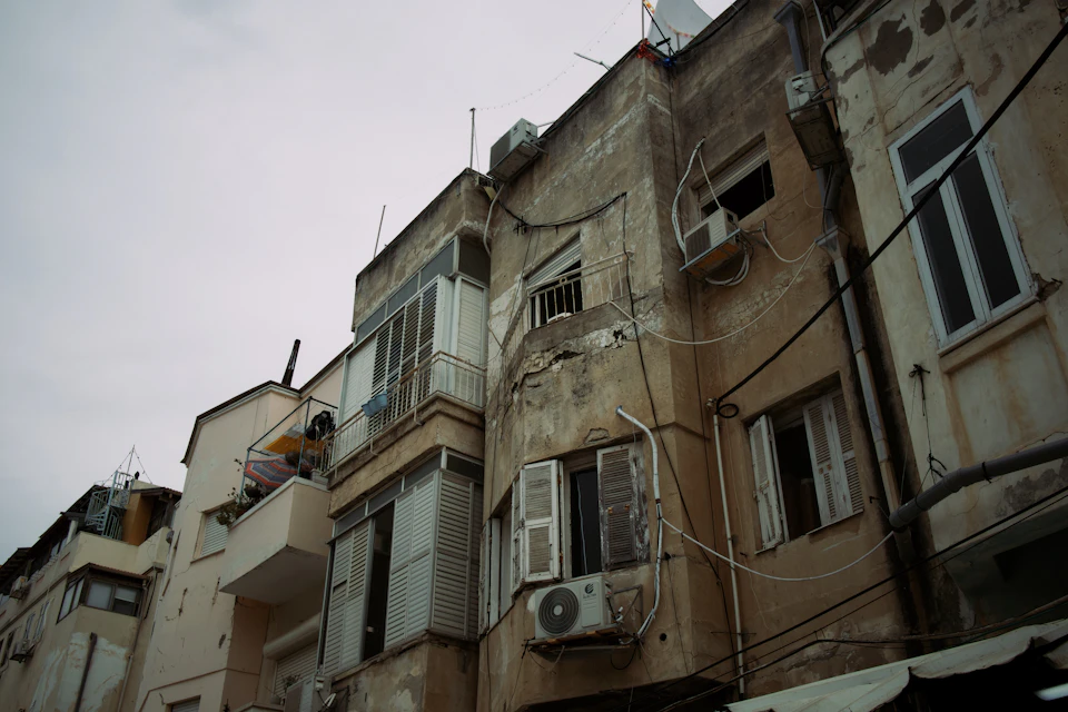 The image depicts a row of old, run-down buildings with peeling paint and boarded up windows. The perspective is from below, looking upwards towards the sky, emphasizing the height of the structures. A few people can be seen in the scene, but they are not the main focus. The colors in the photo are muted, reflecting the age and decay of the buildings.