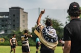 A group of athletes are on a grassy field engaged in a sports activity, likely rugby. One player is seen with a flag draped over their shoulders, pointing upwards. Other players and a referee or coach are visible, all wearing sports uniforms. In the background, there are trees and a building, suggesting an urban setting.