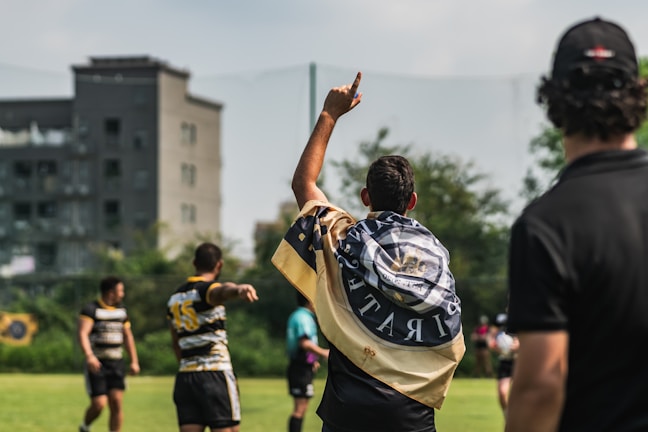 A group of athletes are on a grassy field engaged in a sports activity, likely rugby. One player is seen with a flag draped over their shoulders, pointing upwards. Other players and a referee or coach are visible, all wearing sports uniforms. In the background, there are trees and a building, suggesting an urban setting.