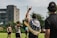 A group of athletes are on a grassy field engaged in a sports activity, likely rugby. One player is seen with a flag draped over their shoulders, pointing upwards. Other players and a referee or coach are visible, all wearing sports uniforms. In the background, there are trees and a building, suggesting an urban setting.