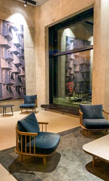 A modern, stylish room with beige concrete walls and a large window reflecting abstract wall art. The space contains wooden chairs with navy blue cushions, placed around a minimalist black round table on a gray textured rug. Warm lighting illuminates the area, enhancing the contemporary aesthetic.
