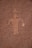 A rock surface features an ancient petroglyph depicting a humanoid figure with outstretched arms. The carving is etched into a textured reddish-brown rock face, typical of prehistoric rock art.
