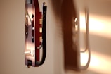 A stringed musical instrument is mounted on a wall. The instrument has a rich wooden body with metal strings and some metallic components. The wall and the instrument are warmly illuminated by sunlight, creating shadows that add depth to the scene.