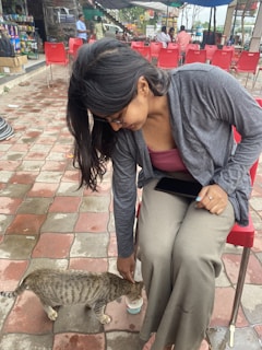 A person with long dark hair and glasses is sitting on a red chair in an outdoor setting. They are wearing a gray cardigan and light brown pants. The person is bending down to feed a tabby cat with spotted fur. The cat is drinking from a small cup placed on the tiled patio ground. Several red chairs and tables are visible in the background, along with a few people and vending machines.