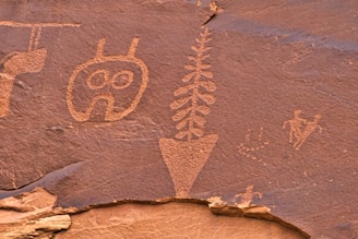 Ancient rock carvings depict various symbols and figures etched into a reddish-brown stone surface. The carvings include geometric shapes, plant-like patterns, and humanoid figures, suggesting symbolic or ritualistic significance.