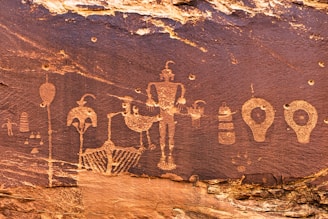 Ancient rock art carvings depict various humanoid and abstract shapes on a stone surface. The carvings include figures with distinct outlines and features, arranged in a sequence across the rock face, showcasing intricate details and patterns.