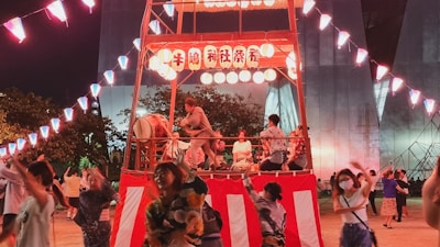 A lively festival scene features people in traditional clothing dancing around a central platform decorated with lanterns. The platform is adorned with red and white stripes and has musicians playing drums. Colorful lights and trees are visible in the background.