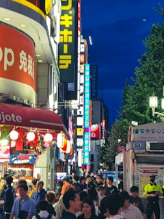 A bustling city street scene at night filled with a crowd of people walking under a canopy of bright neon signs and store lights. The atmosphere is energetic, with a variety of shops lining the street, including a prominently visible store with red signage. Trees are visible on the right side, and a safety station with a security guard is also present.